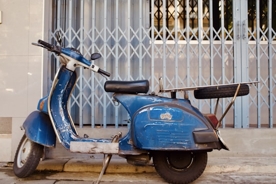 A vintage blue scooter is parked on a concrete surface in front of a metal folding gate. The scooter appears worn, with signs of rust and chipped paint. Mounted on its rear is a metal rack carrying a spare tire.
