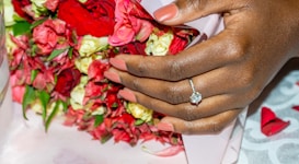 A hand with a manicured style in a soft pink shade is adorned with an elegant diamond ring. The hand clasps a vibrant bouquet of roses, displaying various shades of red, pink, and white. The background has hints of a fabric or textured surface with pinkish hues.