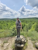 A person wearing outdoor gear and a cap stands on a rocky outcrop surrounded by lush greenery and hills under a partly cloudy sky.