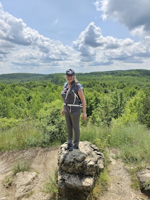 A person wearing outdoor gear and a cap stands on a rocky outcrop surrounded by lush greenery and hills under a partly cloudy sky.