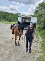 A woman wearing a helmet and equestrian attire stands beside a horse, holding its lead rope. The horse is outfitted with a fly mask and saddle. They are in an open area near a horse trailer, surrounded by greenery and under a partly cloudy sky.