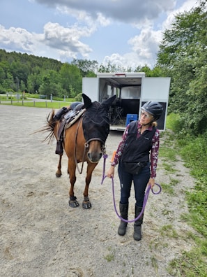 A woman wearing a helmet and equestrian attire stands beside a horse, holding its lead rope. The horse is outfitted with a fly mask and saddle. They are in an open area near a horse trailer, surrounded by greenery and under a partly cloudy sky.