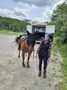 A woman wearing a helmet and equestrian attire stands beside a horse, holding its lead rope. The horse is outfitted with a fly mask and saddle. They are in an open area near a horse trailer, surrounded by greenery and under a partly cloudy sky.