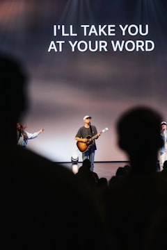 A musician wearing a cap is playing an acoustic guitar on stage with two other visible performers. There is a prominent text above them that reads 'I'LL TAKE YOU AT YOUR WORD'. The stage is well-lit, and silhouettes of a crowd are visible in the foreground.