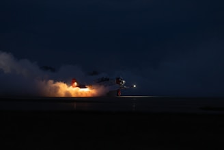 A small aircraft is captured in mid-flight at night, with flames and smoke billowing from the rear, possibly during takeoff or landing. The airplane's lights are illuminated, casting a soft glow on its body.