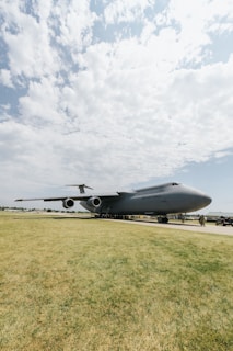 A large military cargo plane is stationed on an airfield. The plane is gray with 'U.S. Air Force' written on the side. The sky is partly cloudy, and several people can be seen near the plane, possibly military personnel.
