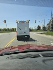 A white service van with a company logo and contact information is stopped at a traffic light. The van is equipped with a ladder on top. It's daytime, with clear skies, and the scene is viewed from inside a red car. Traffic lights and electric poles are visible in the background, surrounded by greenery.