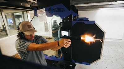A person wearing protective headphones and a cap is firing a pistol at a shooting range. The muzzle flash is visible as the gun discharges. The environment appears to be an indoor shooting facility with safety partitions and a target setup. Another person is visible in the background behind a glass partition.