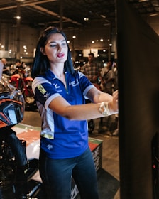 A woman wearing a blue polo shirt with logos is standing indoors, interacting with a large touchscreen or display. She appears to be explaining or demonstrating something. In the background, there are several people and motorcycles in a large, well-lit, industrial-style space.