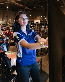 A woman wearing a blue polo shirt with logos is standing indoors, interacting with a large touchscreen or display. She appears to be explaining or demonstrating something. In the background, there are several people and motorcycles in a large, well-lit, industrial-style space.