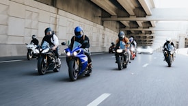 A group of motorcyclists riding together on a multi-lane road under an overpass. All riders are wearing helmets and protective gear. The scene appears to be urban with concrete structures and multiple lanes for traffic. Some cars can be seen in the background.