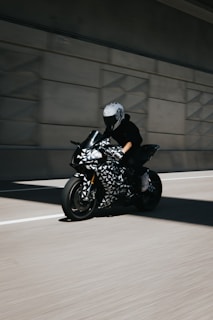 A motorcyclist wearing a helmet rides a sleek motorcycle with a unique black and white geometric pattern on its body. The rider is on a road within a tunnel, creating a dynamic and fast-paced scene.
