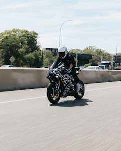 A motorcyclist rides a stylish motorcycle with a geometric black and white pattern on a sunny day. The rider wears a full protective outfit, including a white helmet and black gloves. The background shows a clear sky and trees, suggesting a highway or open road.