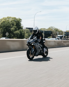 A motorcyclist rides a stylish motorcycle with a geometric black and white pattern on a sunny day. The rider wears a full protective outfit, including a white helmet and black gloves. The background shows a clear sky and trees, suggesting a highway or open road.
