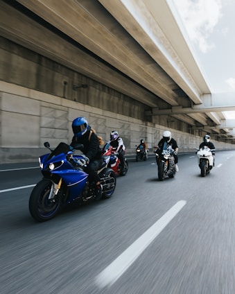 A group of motorcyclists rides along an urban highway. The riders are wearing helmets and protective gear, and the motorcycles are of different colors and styles. The scene is captured from a dynamic angle, emphasizing speed and motion, with the highway bridge overhead adding to the urban setting.