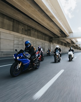 A group of motorcyclists rides along an urban highway. The riders are wearing helmets and protective gear, and the motorcycles are of different colors and styles. The scene is captured from a dynamic angle, emphasizing speed and motion, with the highway bridge overhead adding to the urban setting.