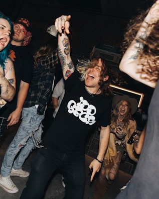 A lively group of people is enjoying a night out, possibly at a club or concert. The scene is vibrant, with individuals laughing, dancing, and showing expressive body gestures. One person with tattoos and colorful hair is at the forefront, energetically pointing and engaging with the moment. The atmosphere is casual and fun.