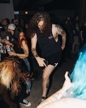 A group of people in a dimly lit room are engaged in a lively gathering. Two individuals with tattoos are noticeably energetic, appearing to dance or enjoy music. The atmosphere suggests a casual, social event with drinks in hand and various expressions of enjoyment. The presence of tattoos and casual attire indicates a youthful, alternative scene.