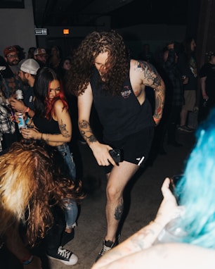 A group of people in a dimly lit room are engaged in a lively gathering. Two individuals with tattoos are noticeably energetic, appearing to dance or enjoy music. The atmosphere suggests a casual, social event with drinks in hand and various expressions of enjoyment. The presence of tattoos and casual attire indicates a youthful, alternative scene.