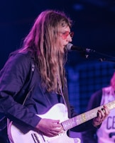A person with long hair and wearing red-tinted glasses is playing an electric guitar and singing into a microphone on stage. The lighting is dim, with some purple and blue shades illuminating the scene. The person is dressed in a dark jacket.
