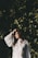 A woman in a white, flowing, pleated blouse stands in front of a wall covered with lush green ivy. She has long, wavy hair and appears to be gazing downwards with a contemplative expression, as sunlight casts soft shadows across her face and the leaves.