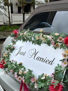 A car is adorned with a floral wreath featuring assorted flowers, predominantly pink and red, surrounding a sign that reads 'Just Married.' The car is parked near an outdoor setting with trees and a white railing in the background.