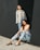 Two women in casual attire pose against a large, dark wall. One woman stands, wearing a light jacket over a top and jeans, looking upward with a contemplative expression. The other woman sits on the ground, leaning back against the wall in a relaxed manner, wearing a white top, jeans, and black sandals. The ground is sandy and bright.