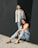 Two women in casual attire pose against a large, dark wall. One woman stands, wearing a light jacket over a top and jeans, looking upward with a contemplative expression. The other woman sits on the ground, leaning back against the wall in a relaxed manner, wearing a white top, jeans, and black sandals. The ground is sandy and bright.