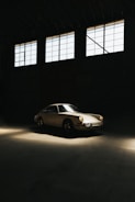 A vintage car is parked inside a dimly lit garage with large windows above. The light casts a warm glow on the sleek body of the car, highlighting its classic design.