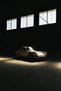 A vintage car is parked inside a dimly lit garage with large windows above. The light casts a warm glow on the sleek body of the car, highlighting its classic design.