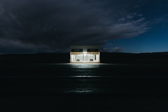 A small, isolated shop illuminated at night against a dark, expansive sky. The shop has a minimalistic design with two large windows displaying designer handbags and shoes. Above the windows, an awning features a brand's name.