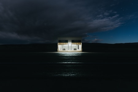 A small, isolated shop illuminated at night against a dark, expansive sky. The shop has a minimalistic design with two large windows displaying designer handbags and shoes. Above the windows, an awning features a brand's name.