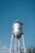 A tall, silver water tower with the word 'MARFA' written on it stands against a clear blue sky, surrounded by some sparse tree branches in the foreground.
