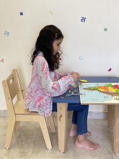 A young girl with long dark hair sits at a small wooden table, focusing intently on assembling a puzzle. She wears blue jeans and a pink and white patterned blouse. Various colorful letters and numbers are scattered on a light-colored wall in the background.