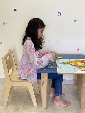 A young girl with long dark hair sits at a small wooden table, focusing intently on assembling a puzzle. She wears blue jeans and a pink and white patterned blouse. Various colorful letters and numbers are scattered on a light-colored wall in the background.