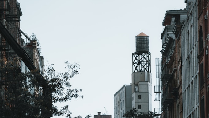 A view of an urban streetscape featuring tall buildings on either side with a prominent water tower on a rooftop. The scene includes a few trees and fire escapes along the sides of the buildings.