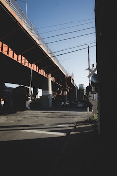 An urban scene with an elevated railway track casting shadows on the street below. The track is painted in orange and supported by concrete pillars. Rail tracks run across the road, with traffic signals and a crossing gate forming part of the infrastructure. Cars are parked in the distance under the bridge.