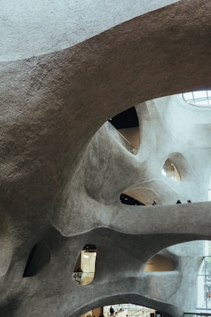 The interior view showcases an architectural structure with organic, flowing curves and textured surfaces. The design features multiple levels and open spaces, with people visible on different floors. The structure allows natural light to enter through the apertures, illuminating the space.