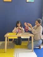 A woman and a young girl are engaged in a crafting activity at a wooden table. The girl is threading beads onto a string, while the woman is doing a similar activity with red beads. They are seated on a yellow and blue patterned floor, against a deep blue wall with two framed pictures partially visible at the top.