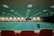 A large indoor sports facility featuring a green playing surface, similar to a bowling green. The ceiling is lined with numerous fluorescent lights, giving the space a bright yet serene atmosphere. Red chairs are arranged along the walls, and a scoreboard can be seen in the distance. A person is actively engaged in the game, bending forward as they release a ball, while inset a few spectators or participants are visible on the left side of the space.