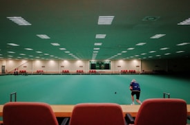 A large indoor sports facility featuring a green playing surface, similar to a bowling green. The ceiling is lined with numerous fluorescent lights, giving the space a bright yet serene atmosphere. Red chairs are arranged along the walls, and a scoreboard can be seen in the distance. A person is actively engaged in the game, bending forward as they release a ball, while inset a few spectators or participants are visible on the left side of the space.