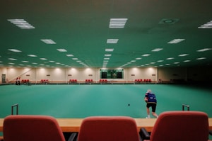 A large indoor sports facility featuring a green playing surface, similar to a bowling green. The ceiling is lined with numerous fluorescent lights, giving the space a bright yet serene atmosphere. Red chairs are arranged along the walls, and a scoreboard can be seen in the distance. A person is actively engaged in the game, bending forward as they release a ball, while inset a few spectators or participants are visible on the left side of the space.