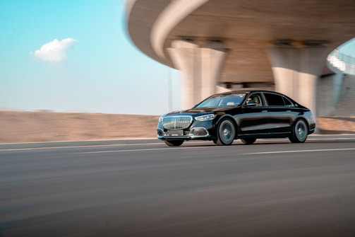A sleek black luxury car driving swiftly on a highway, with an architectural overpass structure in the background. The road is smooth and the sky is clear, suggesting a pleasant day.