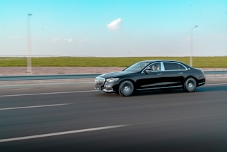 A sleek black luxury car is driving swiftly on a highway with a blurred effect to emphasize motion. The road is smooth and features multiple lanes with clear marking lines. In the background, a strip of green fields contrasts with the bright blue sky, which is dotted with a few white clouds.