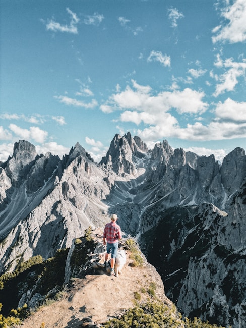 A person in a red plaid shirt and denim shorts stands on a narrow mountain path surrounded by dramatic, jagged peaks. The scene is enhanced by a bright blue sky with scattered clouds, creating a breathtaking alpine vista. A dog accompanies the person, highlighting a sense of companionship and adventure.