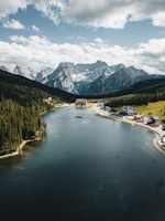 A vast lake is surrounded by dense pine forests and majestic mountains in the background. Several buildings, including hotels or lodges, are situated near the shoreline. The sky is filled with scattered clouds, contributing to the dramatic landscape.
