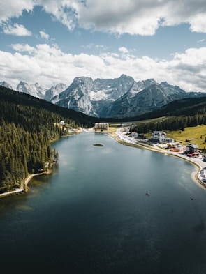 A vast lake is surrounded by dense pine forests and majestic mountains in the background. Several buildings, including hotels or lodges, are situated near the shoreline. The sky is filled with scattered clouds, contributing to the dramatic landscape.