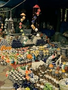 A market stall displaying an array of vibrant ceramic items, including mugs, pots, and decorative figurines. The arrangement includes various colors and patterns, with items neatly organized on tables. Overhead, colorful ceramic wind chimes hang, adding to the overall aesthetic.