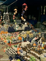 A market stall displaying an array of vibrant ceramic items, including mugs, pots, and decorative figurines. The arrangement includes various colors and patterns, with items neatly organized on tables. Overhead, colorful ceramic wind chimes hang, adding to the overall aesthetic.