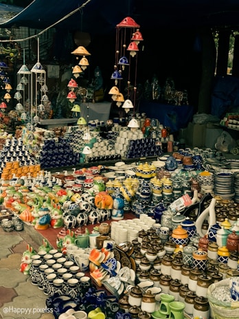 A market stall displaying an array of vibrant ceramic items, including mugs, pots, and decorative figurines. The arrangement includes various colors and patterns, with items neatly organized on tables. Overhead, colorful ceramic wind chimes hang, adding to the overall aesthetic.